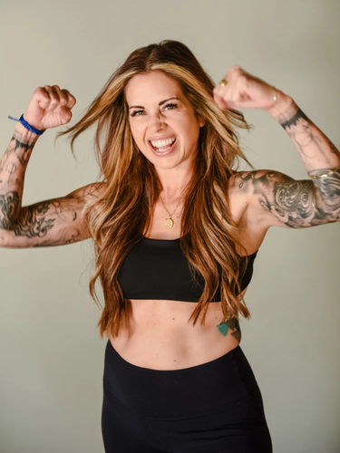 A smiling person with long, crimped hair and full-sleeve tattoos flexes their arms in a celebratory pose.