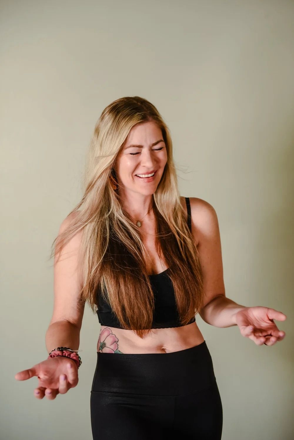 A smiling person with long blonde hair, wearing a black crop top and leggings, gesturing with both hands.