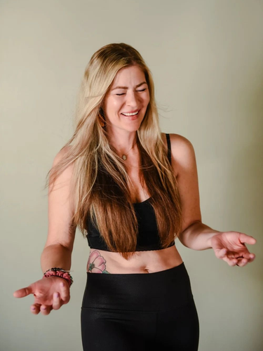 A smiling person with long blonde hair, wearing a black crop top and leggings, gesturing with both hands.