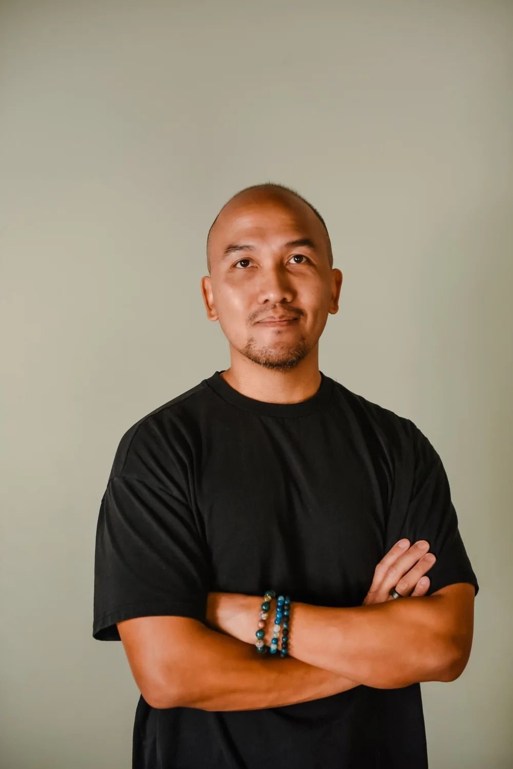A person with a shaved head wearing a black t-shirt and bracelets stands with arms crossed against a neutral background.