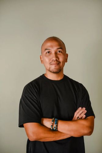 A person with a shaved head wearing a black t-shirt and bracelets stands with arms crossed against a neutral background.