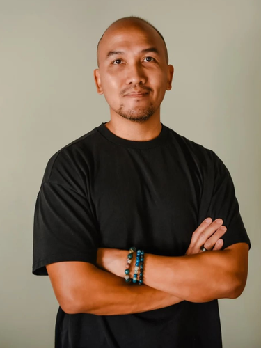 A person with a shaved head wearing a black t-shirt and bracelets stands with arms crossed against a neutral background.
