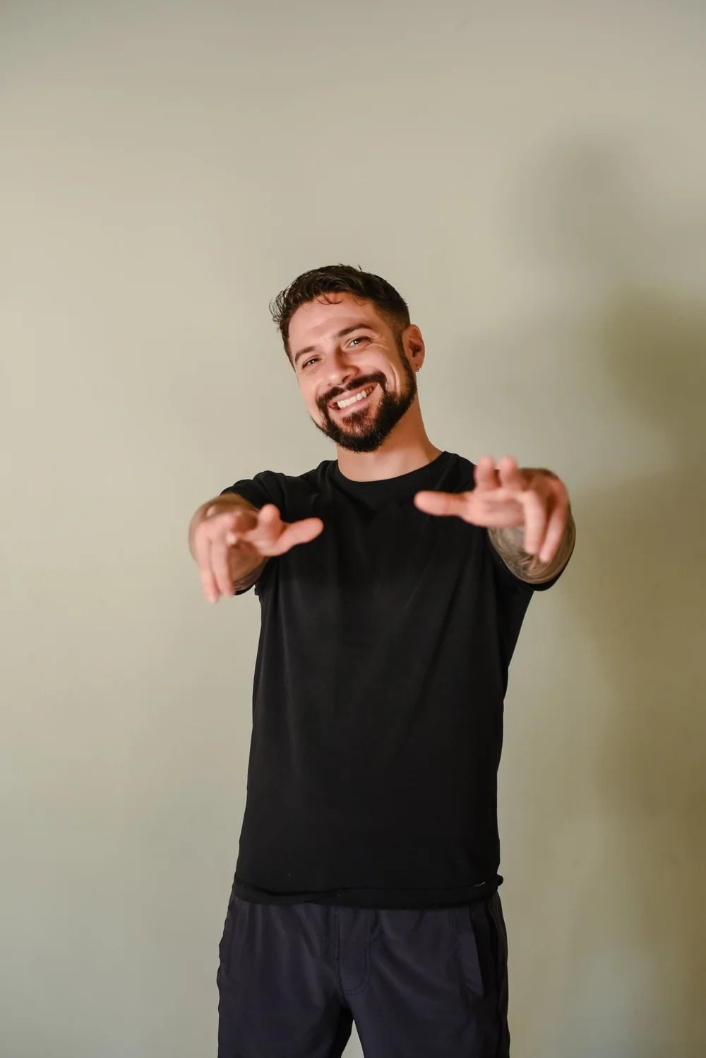 A smiling person with a beard, wearing a black t-shirt and dark shorts, reaching toward the camera against a gray wall.