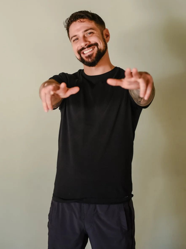 A smiling person with a beard, wearing a black t-shirt and dark shorts, reaching toward the camera against a gray wall.