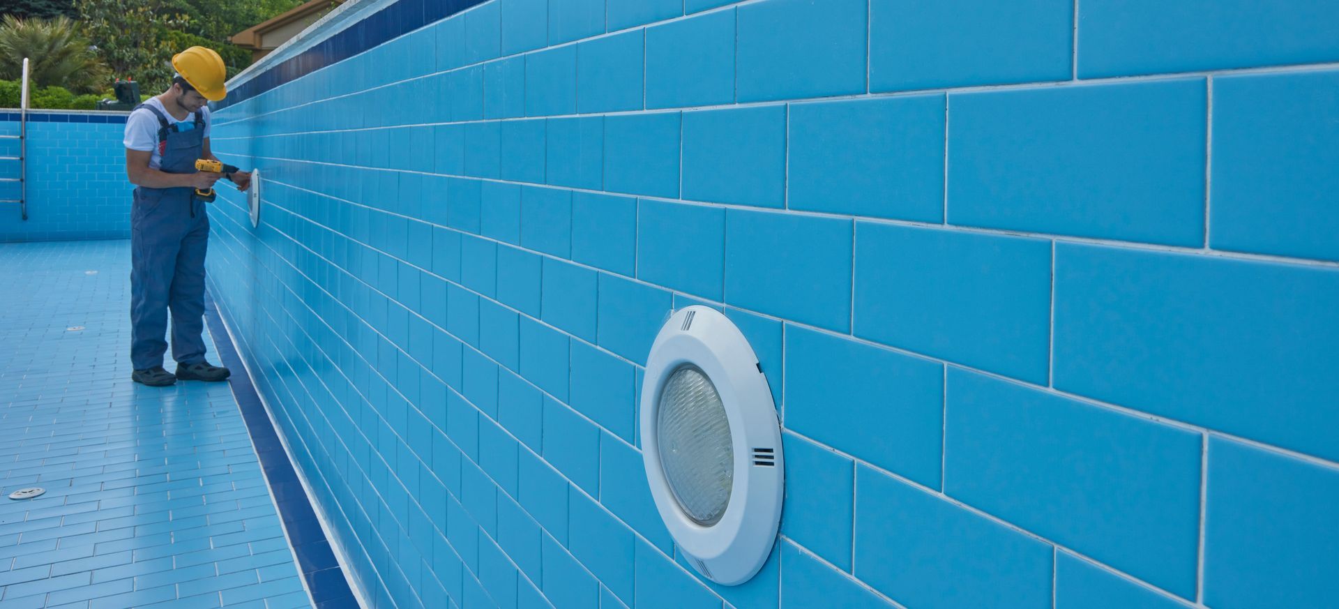 Worker installing blue tile on an empty inground pool wall.