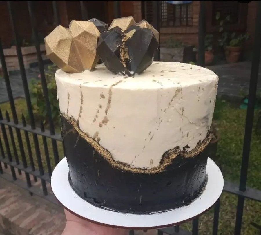 Pastel con glaseado dorado y negro, cubierto con decoraciones geométricas en forma de corazón sobre un plato blanco.