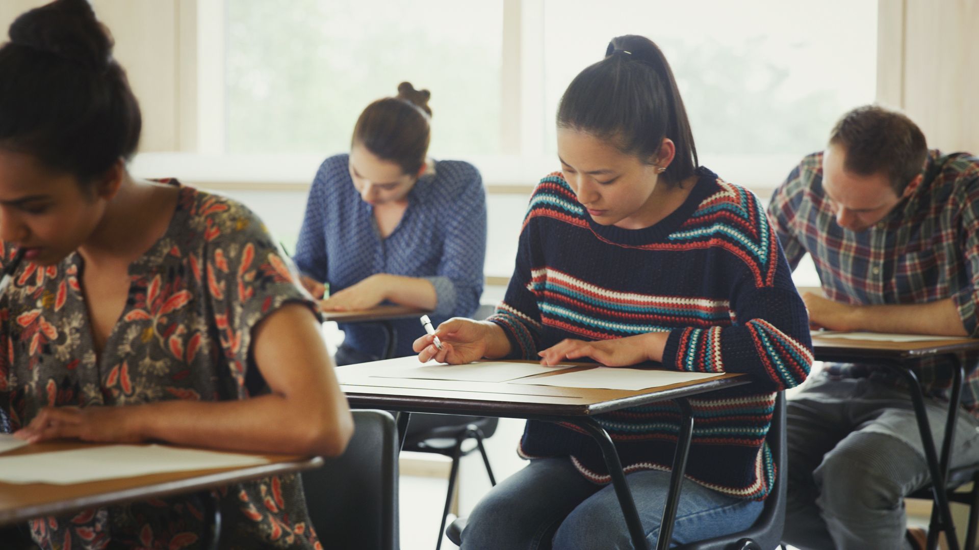 Des personnes passent un examen dans une salle de classe ; focus sur une femme en chemise rayée.