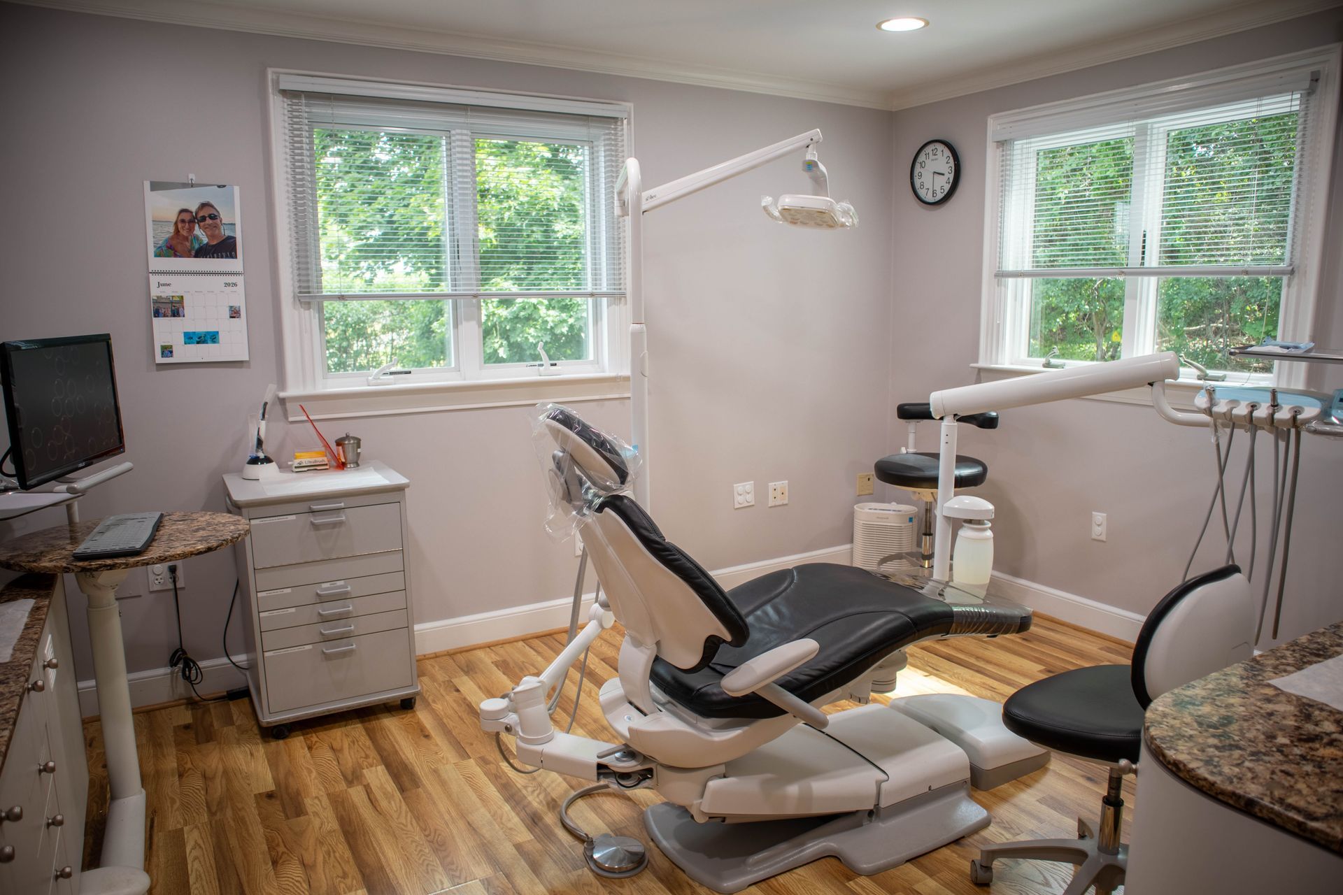 Dental exam room with chair, overhead light, and cabinets by windows in a bright, tidy office