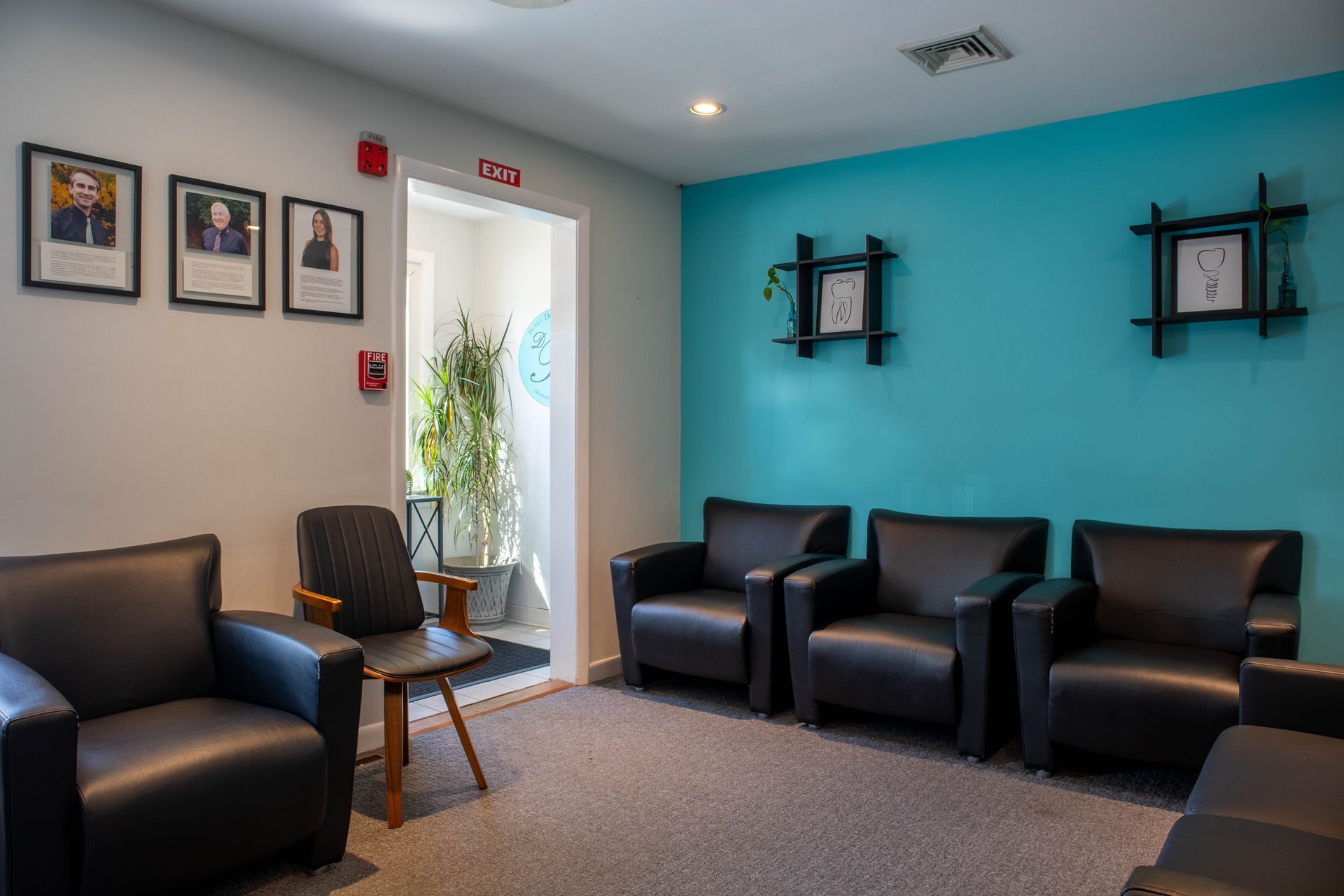 Empty waiting room with black chairs, teal walls, framed photos, and a doorway with daylight.