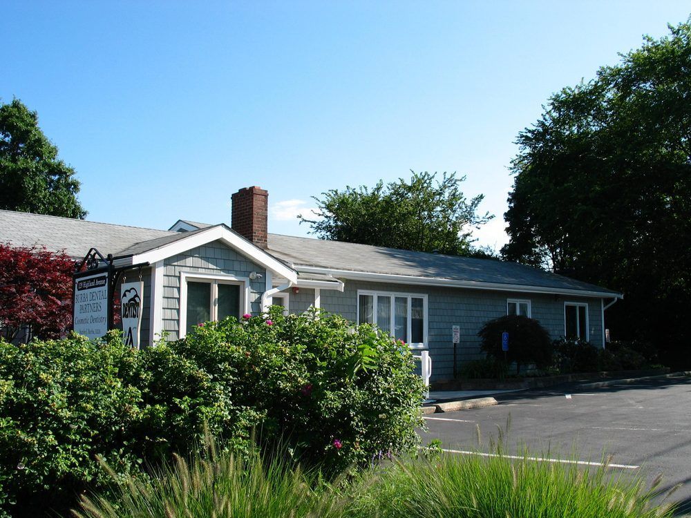 Small gray house with a brick chimney beside a parking lot, surrounded by trees and bushes under a blue sky