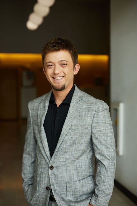Man in a gray blazer and black shirt standing indoors, smiling at the camera