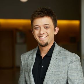Smiling person in a gray blazer and black shirt standing indoors with warm lighting