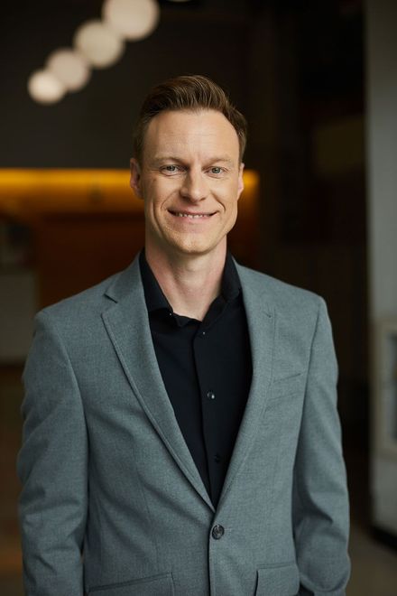 Man in a gray blazer standing indoors, smiling in a softly lit hallway
