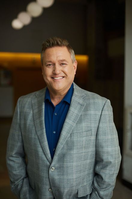 Smiling man in a light gray blazer and navy shirt standing indoors against a blurred dark background