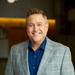 Smiling person in a gray plaid blazer and blue shirt, standing indoors with a blurred warm-lit background