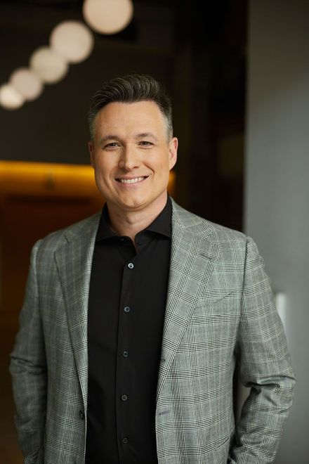 Man in a patterned gray blazer and black shirt smiling indoors against a blurred background