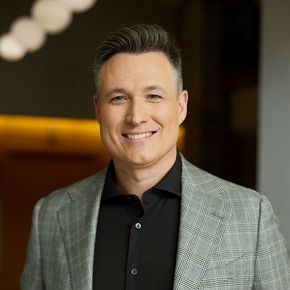 Smiling person in a gray blazer and black shirt against a warm indoor background