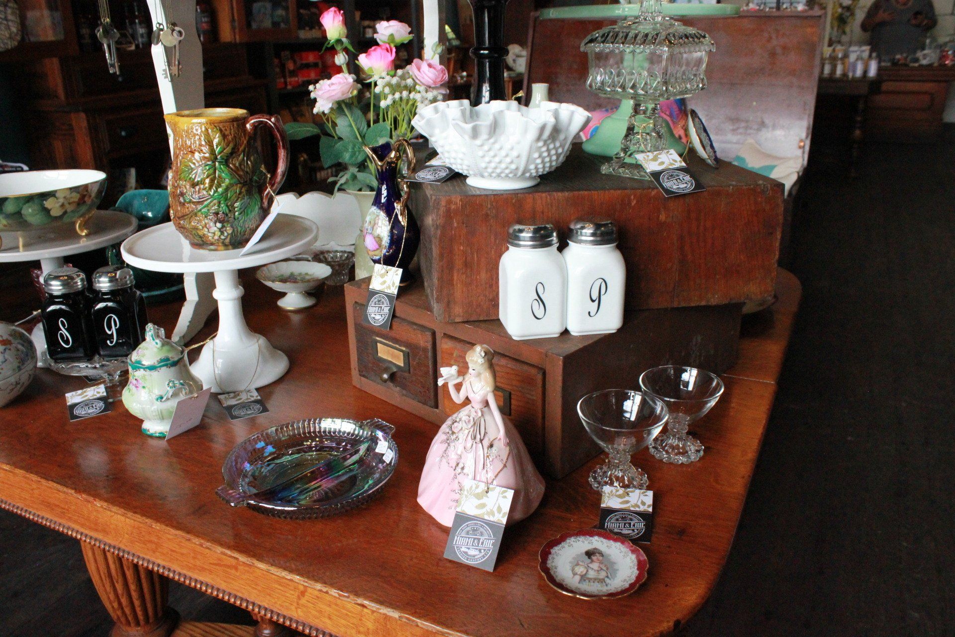 Wooden table displaying vintage dishes, decorations, and a doll in a shop.