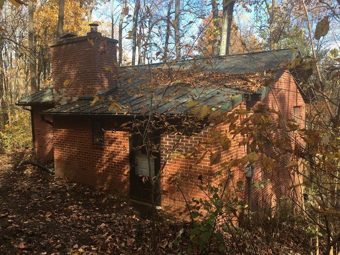 A small brick house in the middle of a forest surrounded by trees and leaves.