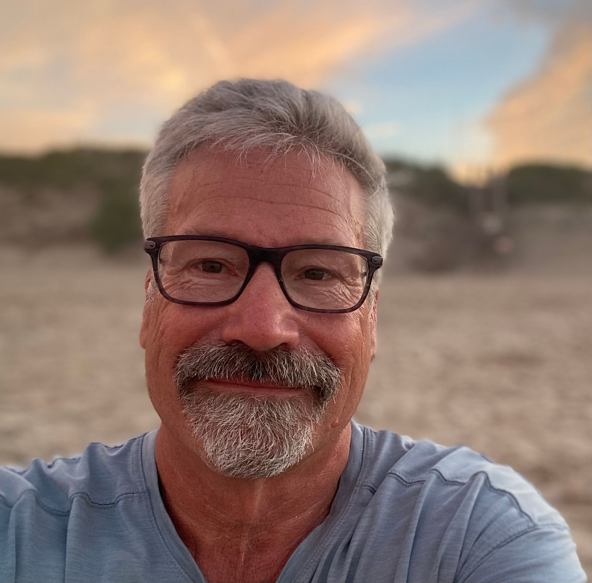 A man with glasses and a beard is taking a selfie on the beach.