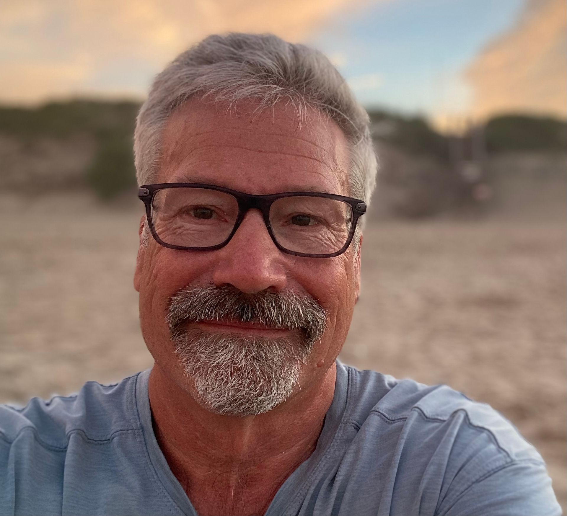 A man with glasses and a beard is taking a selfie on the beach.