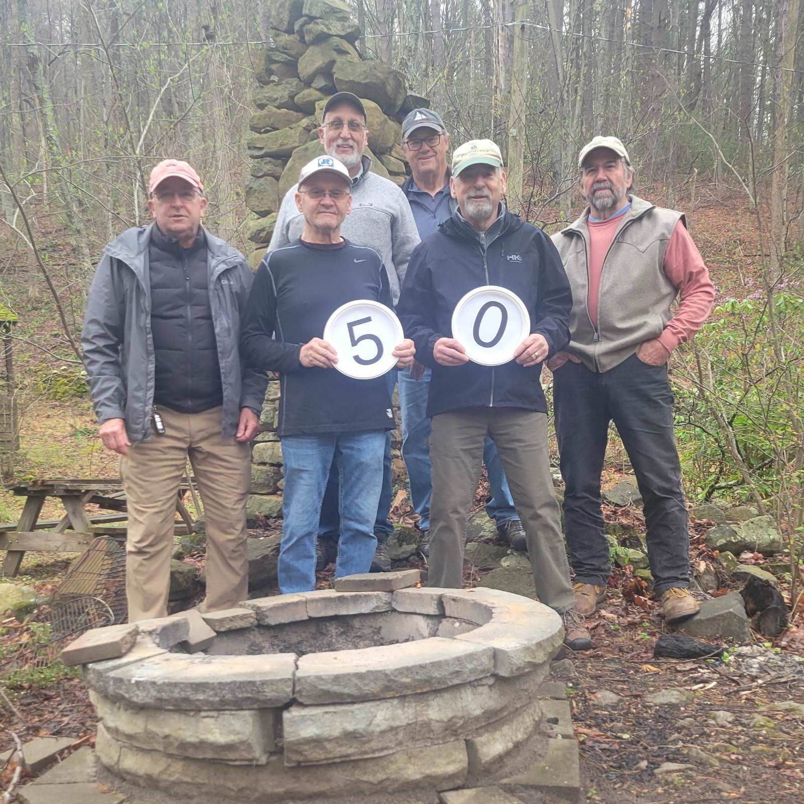 A group of men standing around a fire pit holding plates with the number 50 on them