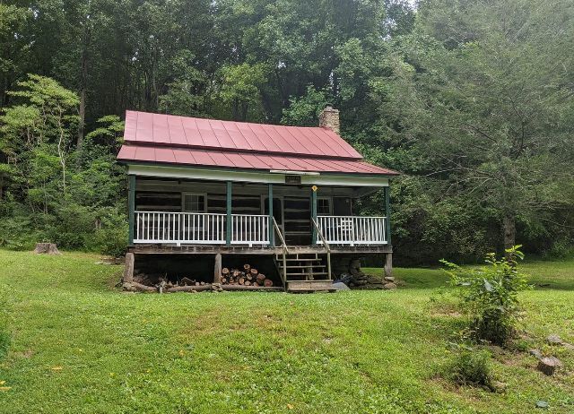A small house with a red roof and a porch in the middle of a grassy field.