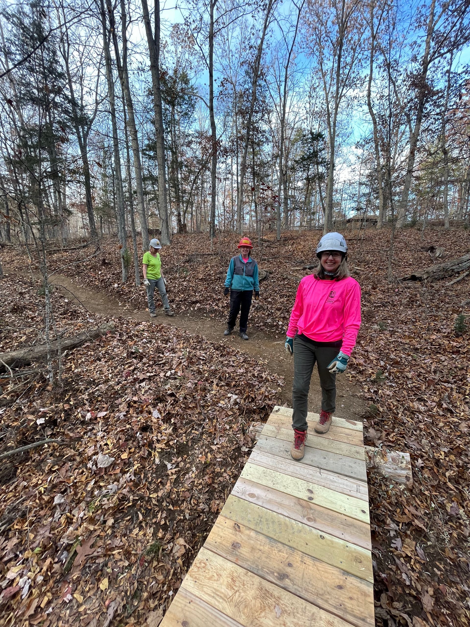 A woman in a pink jacket is standing on a wooden bridge in the woods.