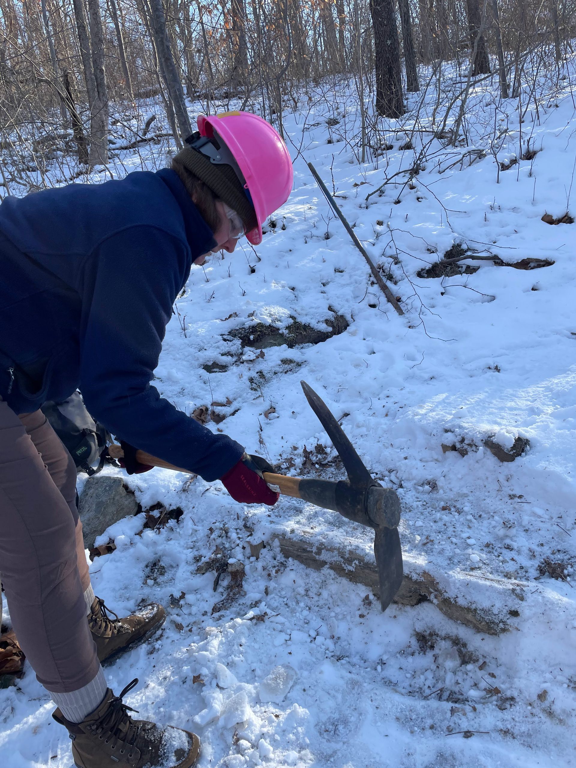 A woman in a pink hard hat is digging in the snow with a pickaxe.