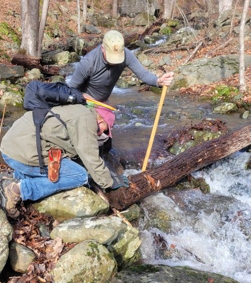 A man is kneeling down next to a stream holding a stick.