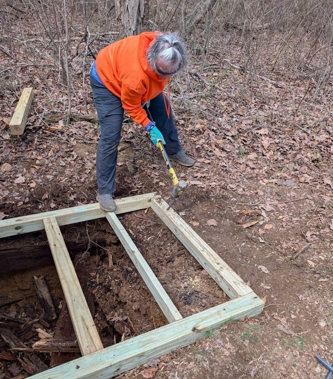 A woman is working on a wooden frame in the woods.