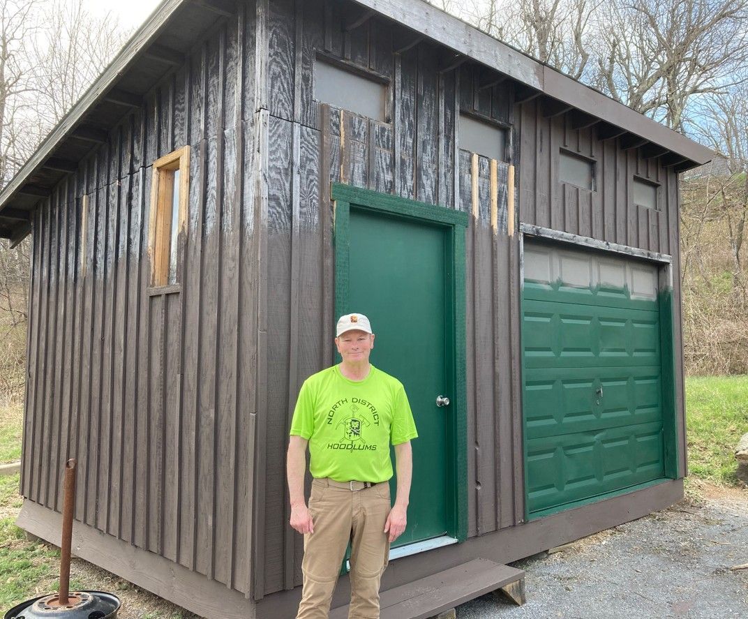 A man in a green shirt is standing in front of a shed.