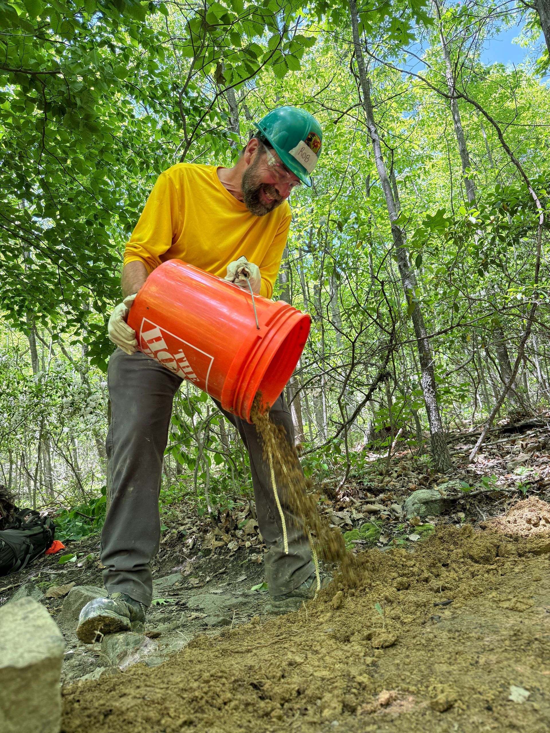 A man is pouring dirt from an orange bucket into a pile.