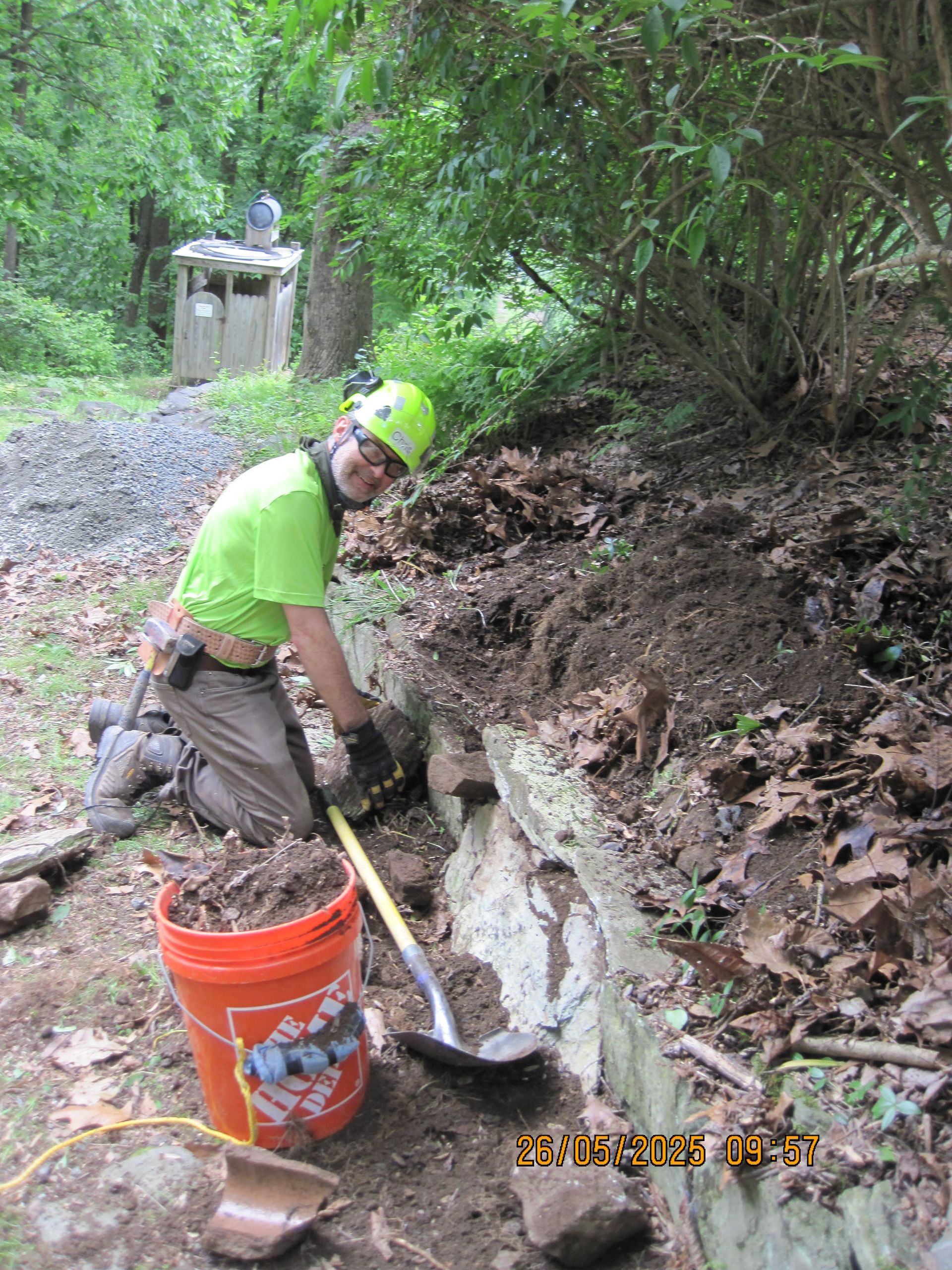 A man is kneeling down next to a bucket of dirt and a shovel.