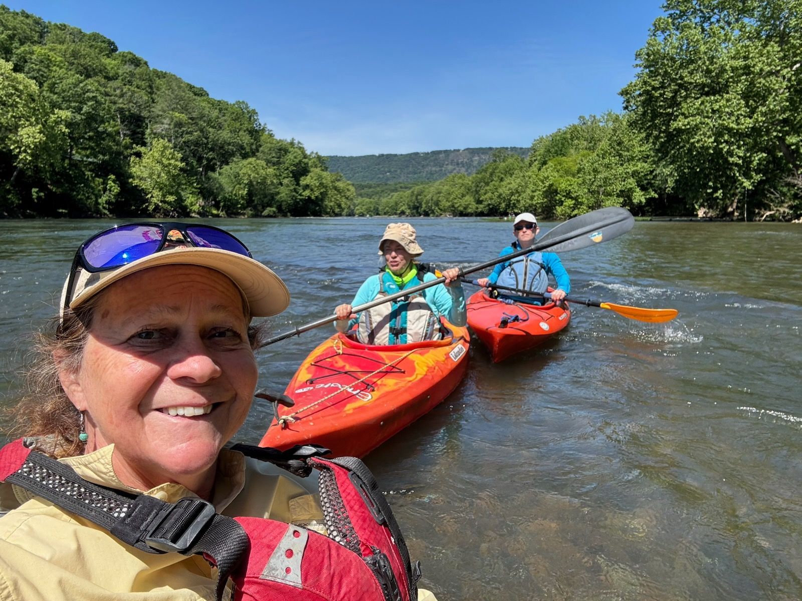 A woman is taking a selfie with two people in kayaks on a river.