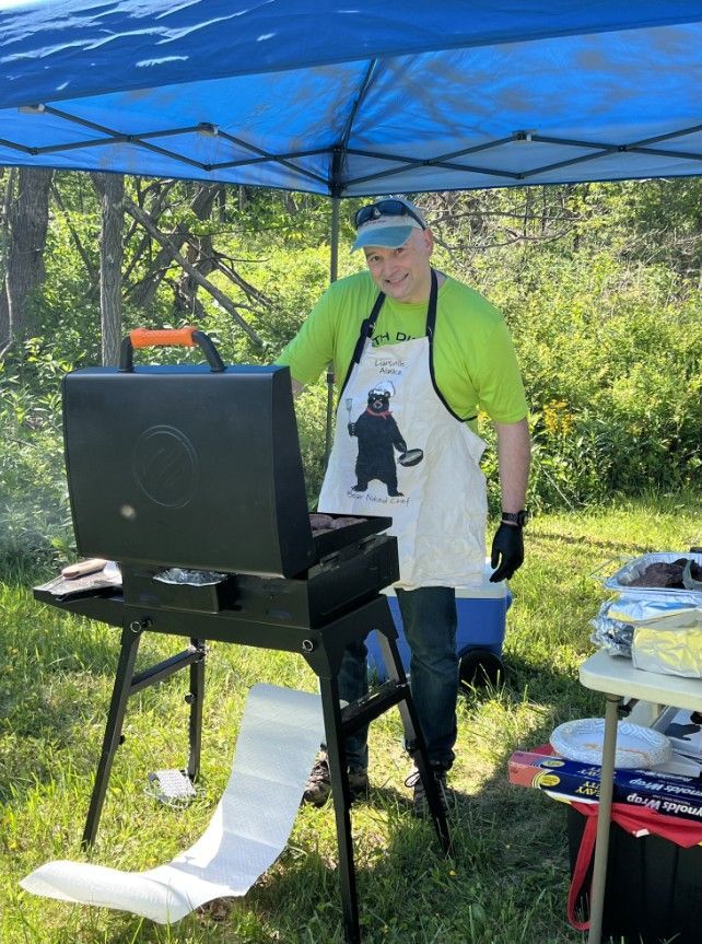 A man wearing an apron with a bear on it is standing in front of a grill.