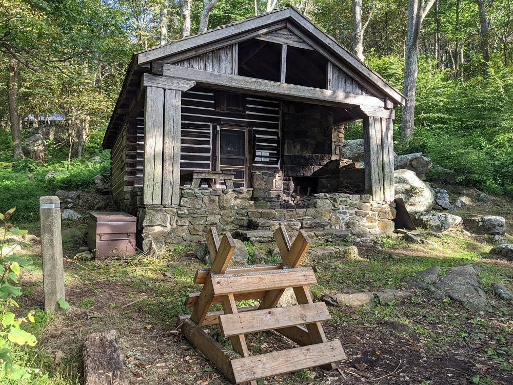 A small log cabin in the middle of a forest with a wooden fence in front of it.