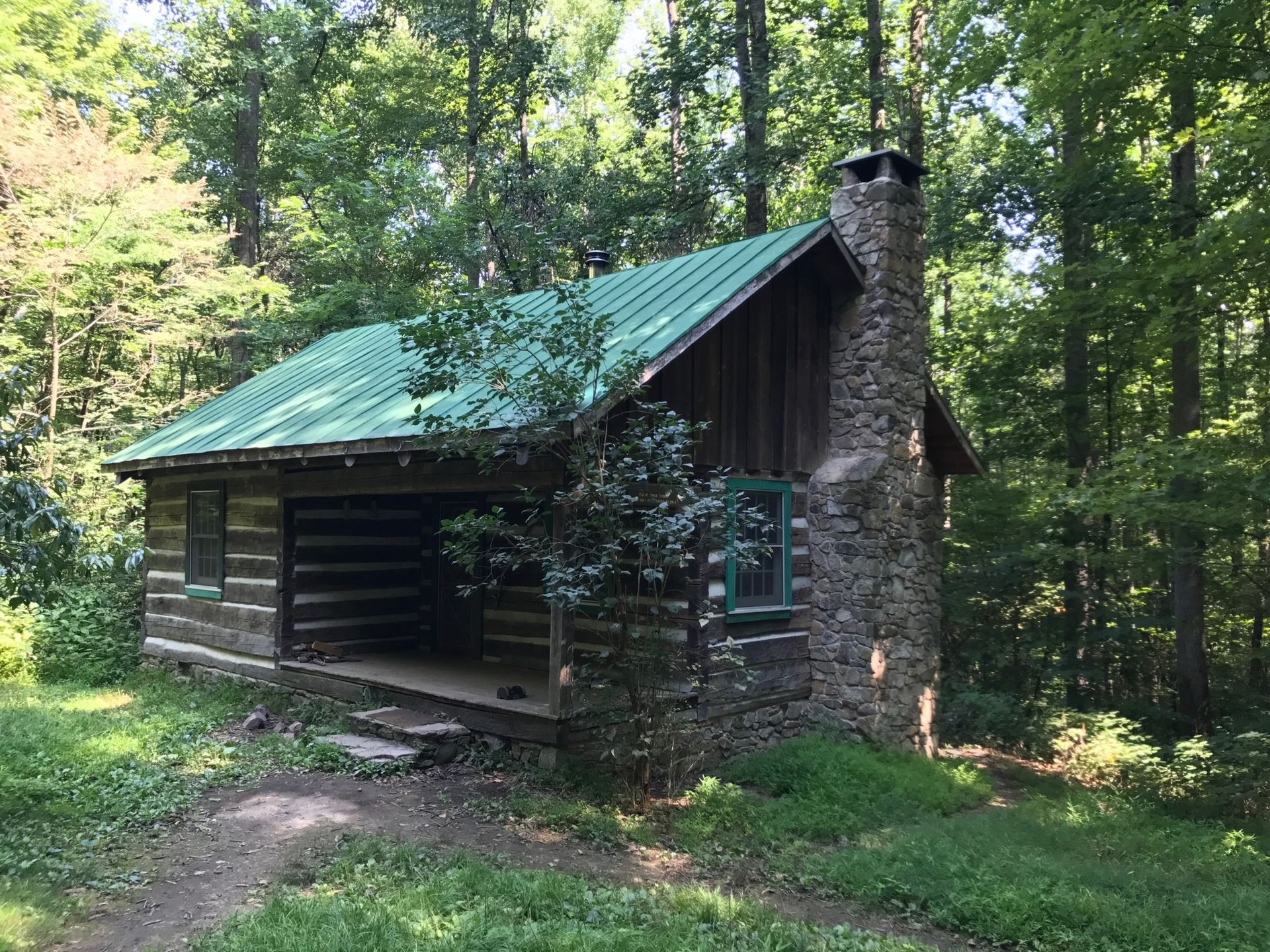 A log cabin with a green roof and a stone chimney in the middle of a forest.