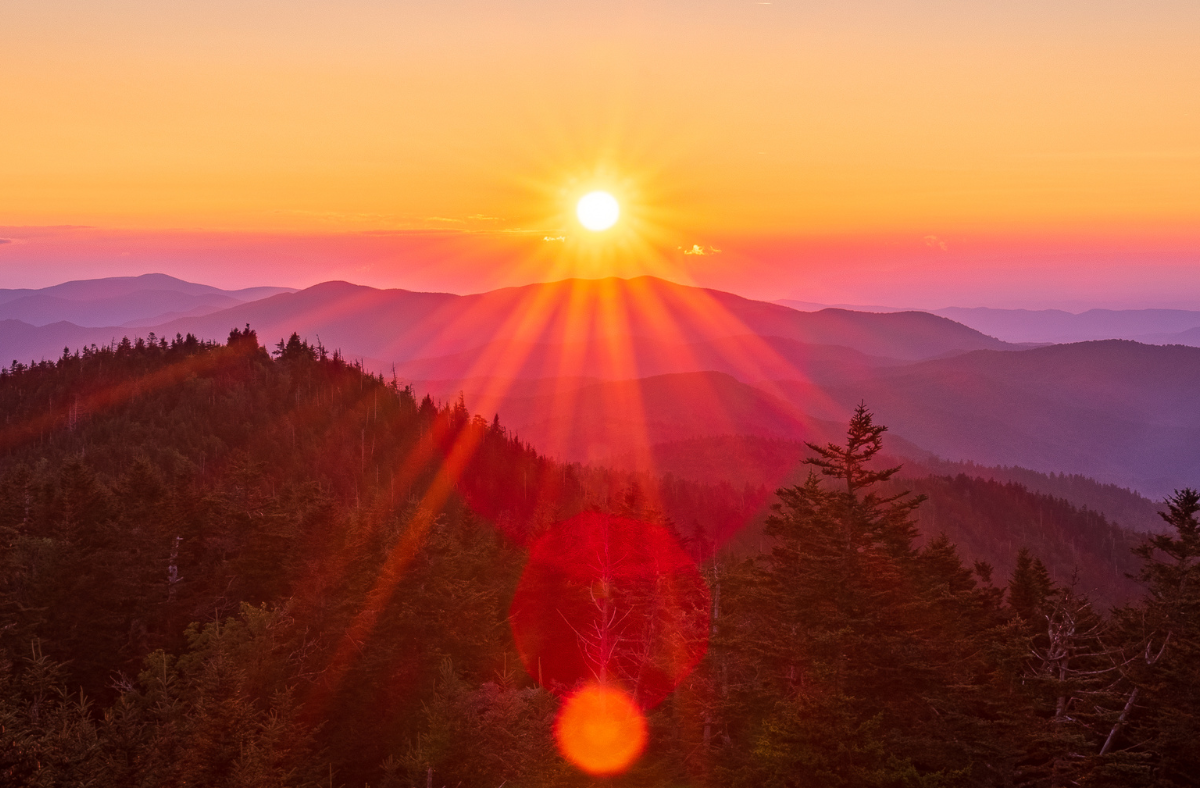 The sun is setting over a mountain range with trees in the foreground.