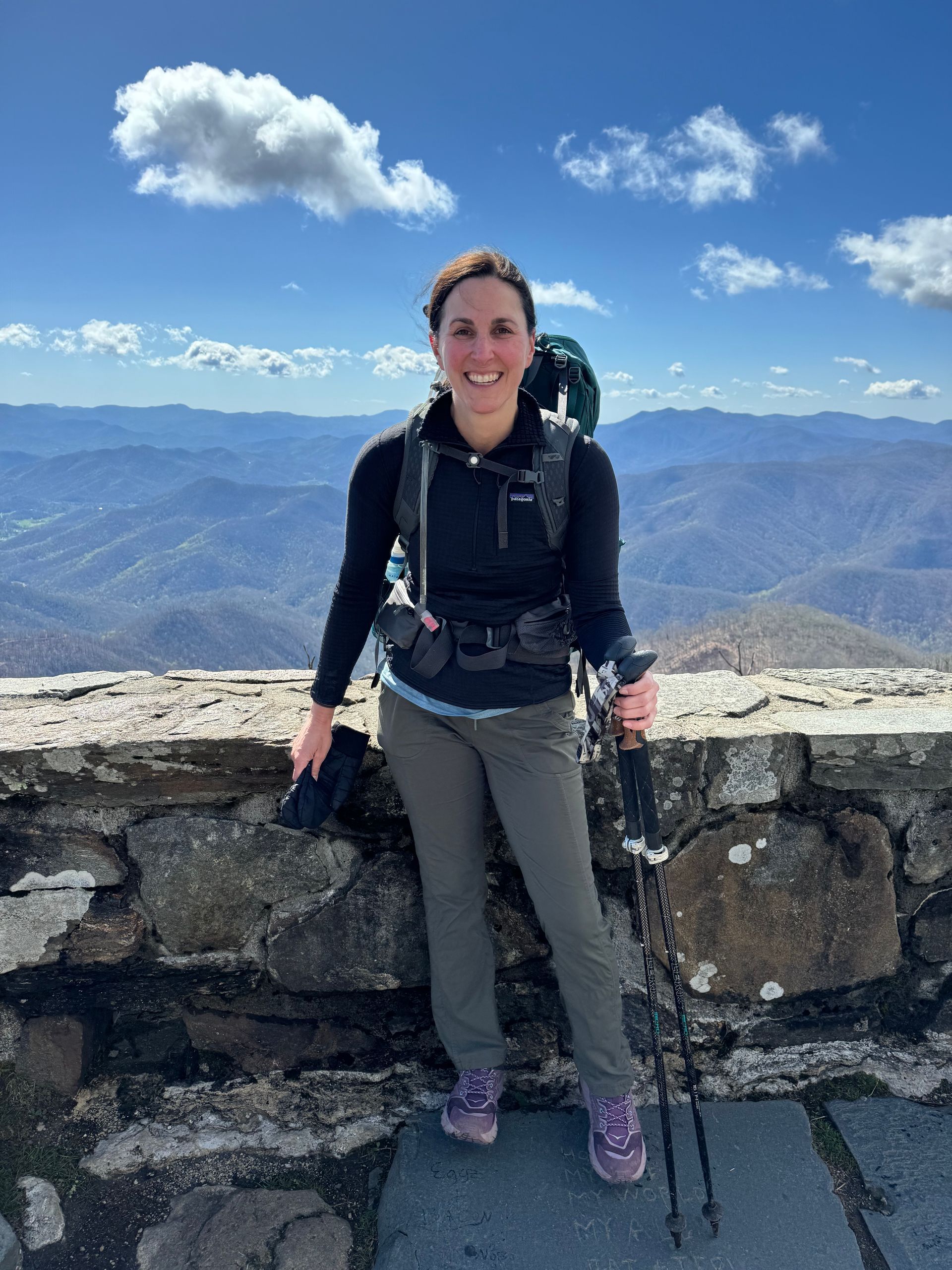 A woman with a backpack and hiking poles is standing on top of a mountain.