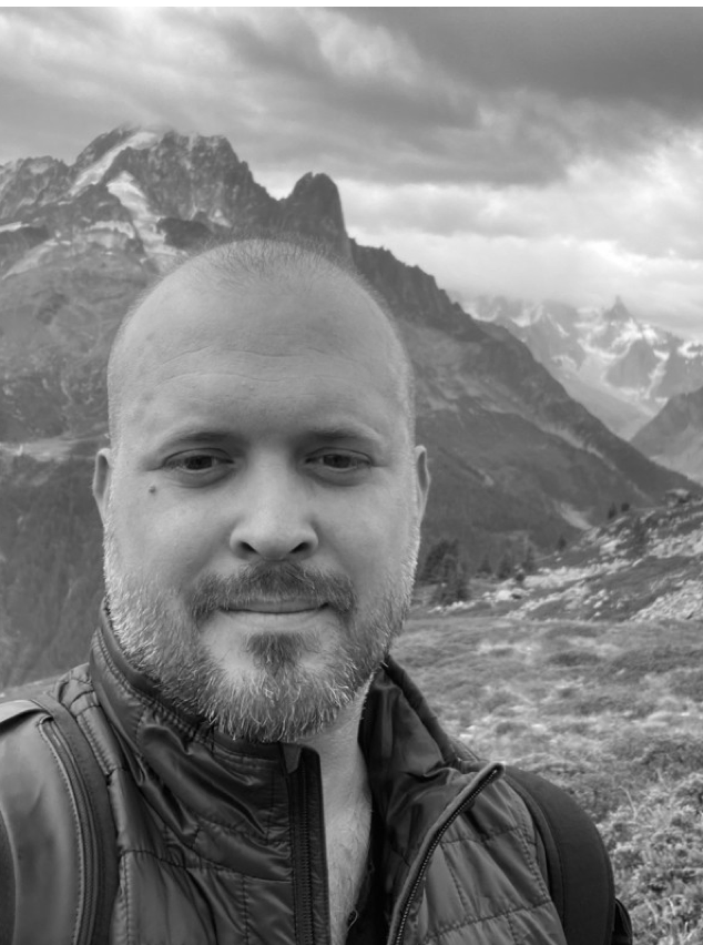 A man with a beard is standing in front of a mountain in a black and white photo.