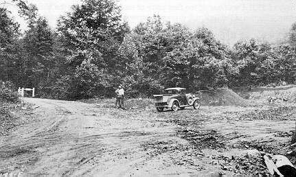 A black and white photo of a man standing next to a car on a dirt road.
