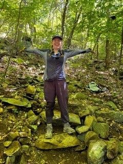 A woman is standing on a pile of rocks in the woods with her arms outstretched.
