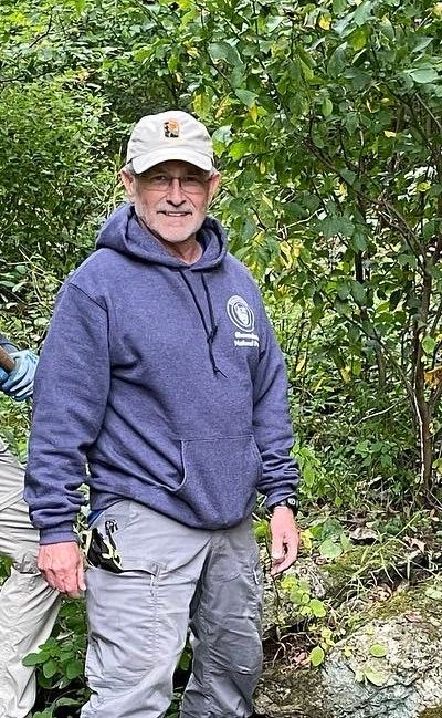 A man in a hoodie and hat is standing in the woods.