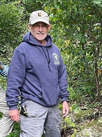 A man in a blue hoodie and hat is standing in the woods.