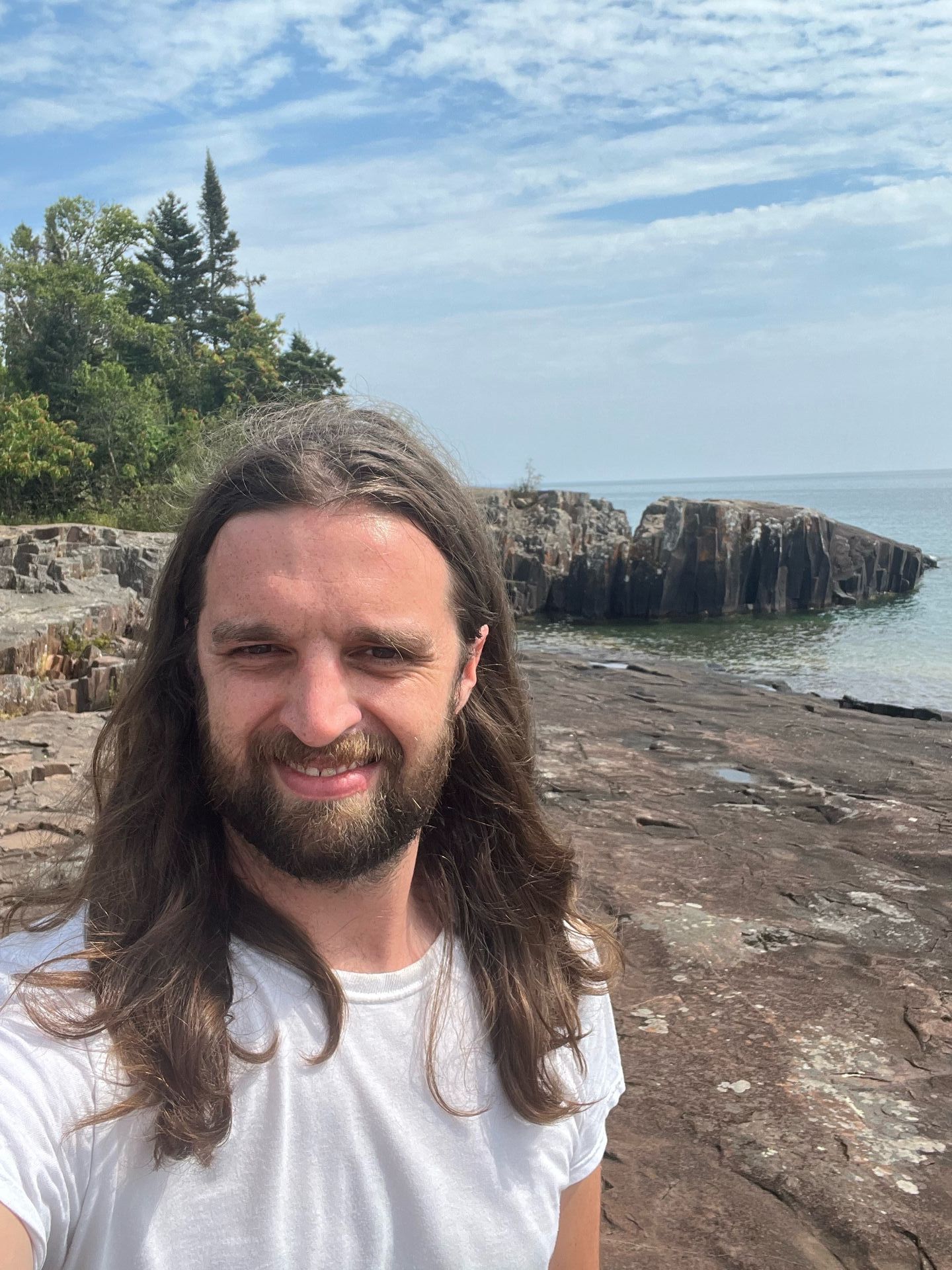 A man with long hair and a beard is standing on a rocky beach.