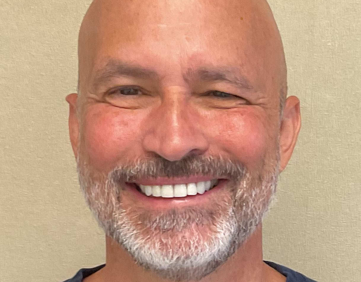 A person with a shaved head and a light gray beard smiling at the camera against a neutral, beige background.