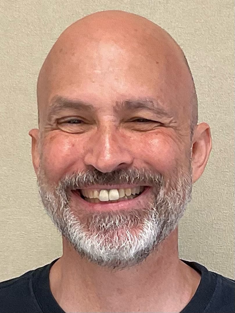 A smiling person with a shaved head and a gray-flecked beard against a plain, light-colored background.