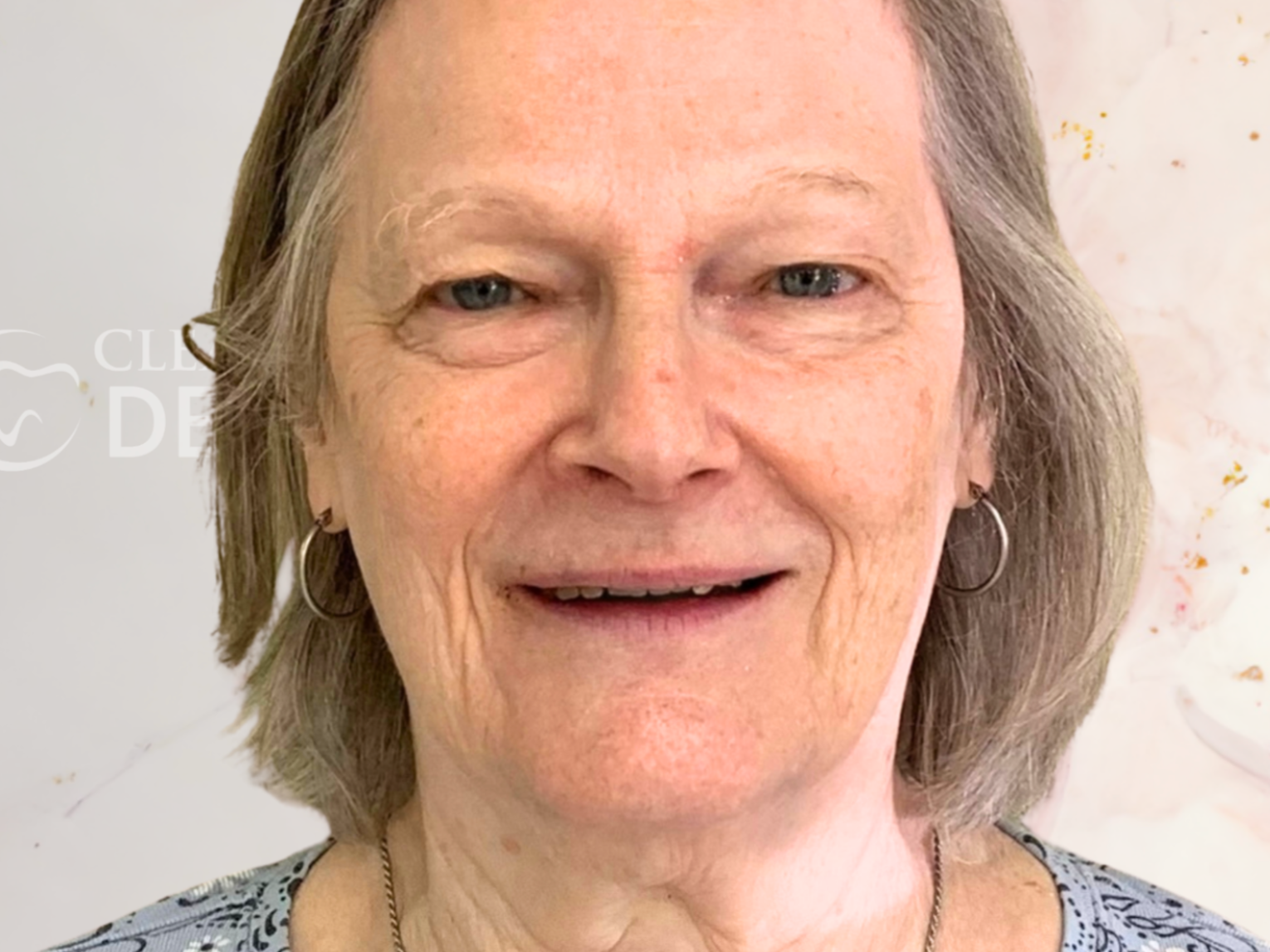 A person with shoulder-length gray hair wearing silver hoop earrings and a patterned top, looking toward the camera.