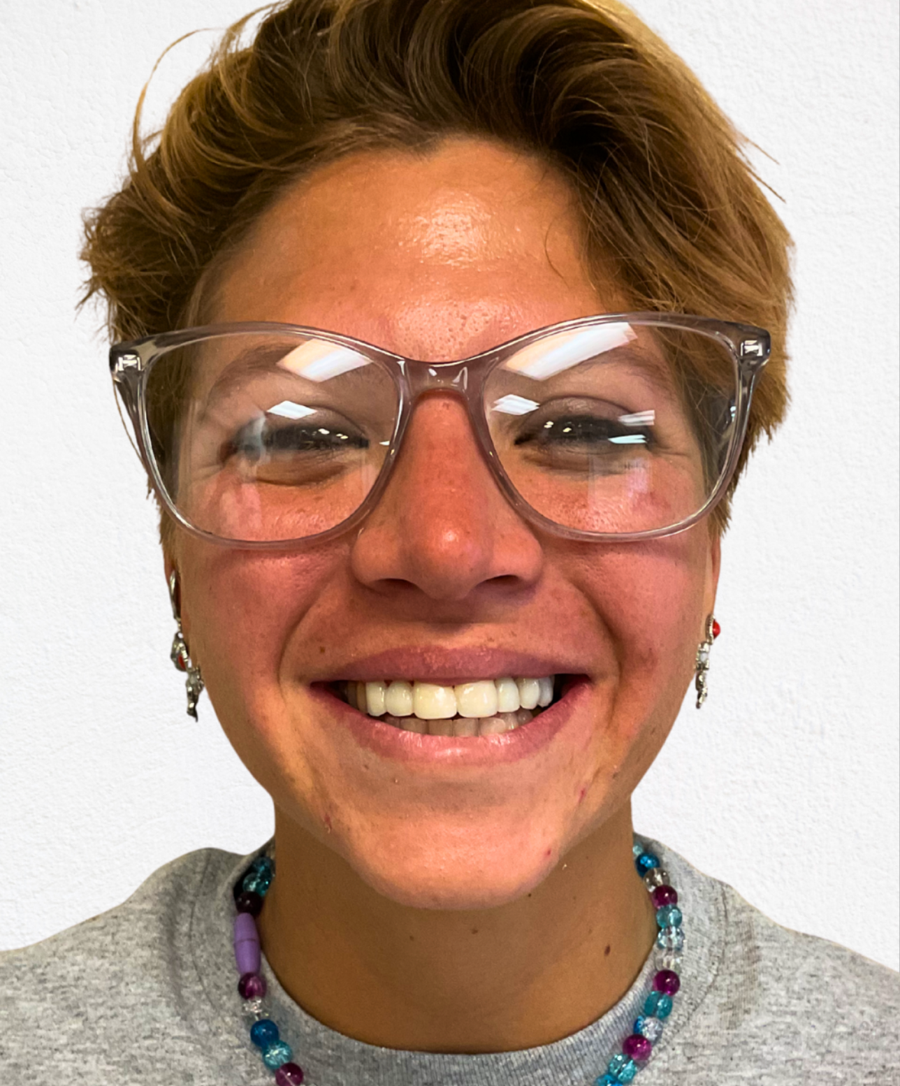 A person with short light-brown hair and clear-framed glasses smiling against a plain white background.